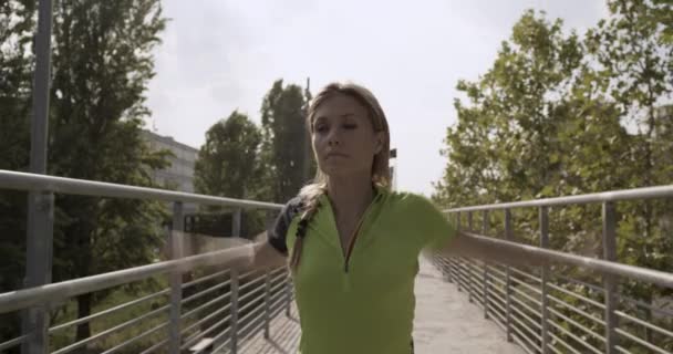 Νεαρή ξανθιά γυναίκα στο φορούν γυμναστήριο προθέρμανση διατάσεις για την εκτέλεση του κατάρτιση. Ηλιόλουστη μέρα του καλοκαιριού. Πράσινη βιομηχανική πόλη. Αστικό δρομέα καρδιο υγιή δραστηριότητα workout.4k αργή κίνηση 60p βίντεο — Αρχείο Βίντεο