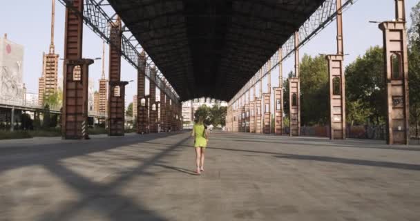 Mujer rubia joven en entrenamiento de correr ropa de fitness en el área industrial.Volver siguiente view.Summer día soleado. ciudad verde industrial.Corredor urbano ejercicio de actividad cardio saludable.4k cámara lenta 60p video — Vídeo de stock