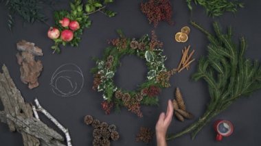 Genel gider kişi eller çelenk garland hazırlanıyor. Doğal yaprakları, çam kozalakları, ağaç kabuğu, tarçın, portakal, nar ile karanlık tablonun dikey üstten görünüm. Xmas tatil sezonu sosyal kartı background.4k video
