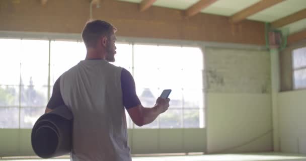 Jonge volwassen man lopen met glooiende mat gebruikend smartphone voor fitness sport training. Grunge industriële stedelijke training.4k slowmotion video — Stockvideo