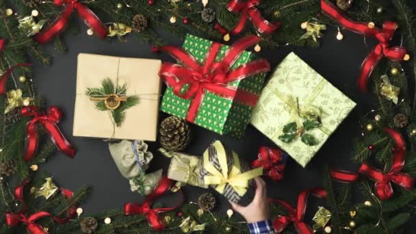 Overhead hand putting gift box with other wraps on dark gray background with decoration frame of pine cones,red and gold ribbons.Vertical top view.Xmas holiday season social card.4k video — Stock Video