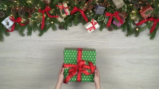 Manos arriba poniendo caja de regalo.Marco de decoración sobre fondo de madera clara con conos de pino, cintas rojas, luces parpadeantes.Espacio de copia de texto o logotipo. — Vídeo de stock