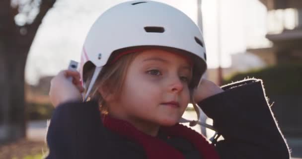 Happy daughter child girl putting on safety helmet before riding bicycle in city park.Childhood, active safety concepts.Sidewalk urban outdoor.Warm sunset cold weather backlight.4k slow motion video — Stock Video