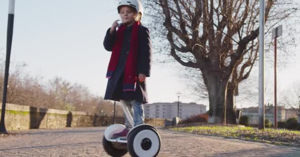 Happy smiling daughter child girl wearing safety helmet portrait riding segway at city park.Childhood, active, safety, future technology.Sidewalk urban outdoor.Warm sunset backlight.4k slow motion video — стоковое видео