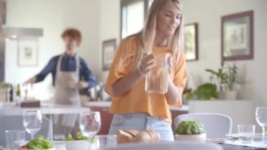 Genç bir kadın yemek için masayı hazırlıyor. Mutlu arkadaşlar konuşurken sürahi koyuyor. Mutlu insanlar modern ev yemeği ya da öğle yemeği partilerinin tadını çıkarıyor.