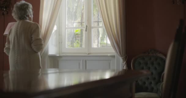 Starší Babičko, Procházka kolem okna a dívá se ven. Krásný bílý vlas starší babička doma. 4k pomalý pohyb videa — Stock video