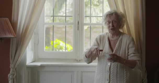 Женщины разных поколений разговаривают вместе. Старшая бабушка улыбается, когда ее внучка приходит к окну и пьет чай или кофе. — стоковое видео