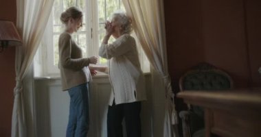 Çok nesilli kadınlar birlikte sarılmayı sever. Büyük büyükanne kadın pencereyakınında torunu veya genç arkadaşı ile mutlu. Evde beyaz saçlı yaşlı büyükanne. Sevgi,birliktelik.4k yavaş hareket