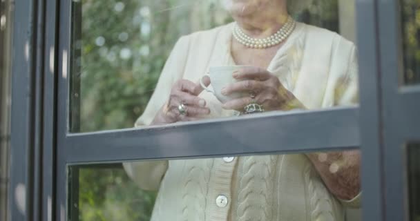 Senior abuela mujer beber taza de té o café cerca de la ventana mirando fuera preocupado o triste o thinking.Beautiful pelo blanco anciano abuela en home.View desde el exterior.4k video en cámara lenta — Vídeos de Stock