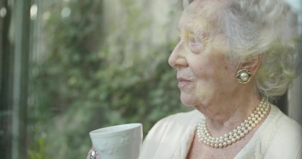 창 가에서 차 나 커피를 마시는 할머니 할머니가 걱정이나 슬픔이나 생각을 하고 있습니다. 집에서 아름 다운 흰 머리 노인 할머니입니다. 외부에서 보기.4k 슬로우 모션 비디오 — 비디오