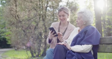 kadın ve üst düzey büyükanne parkta akıllı telefon cihazı kullanarak. Torunu ve büyükanne mobil ile birlikte konuşuyor. Aktif, sevecen, sevgi dolu insan ilişkisi.slow motion video
