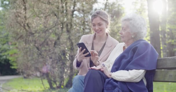 Жінка і старша бабуся, використовуючи пристрій смартфона в парку. Бабуся і бабуся розмовляють разом з мобільним. Активні, турботливі, люблять відносини людей.повільне відео руху — стокове відео