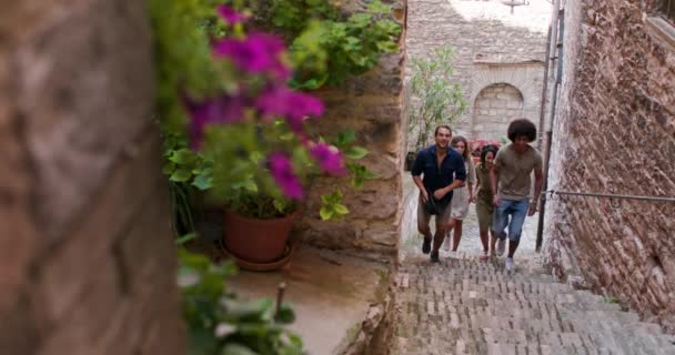 Τέσσερις χαρούμενοι τουριστικοί άνθρωποι περπατούν και τρέχουν μέσα από μικρές σκάλες δρόμου επισκέπτονται την αγροτική πόλη του Spello. Front ακολουθούν. Φίλοι ιταλική εκδρομή για διακοπές στην Ούμπρια. 4K αργή κίνηση — Αρχείο Βίντεο