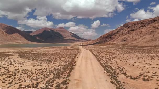 晴れた日に乾燥した砂漠の山の近くの道路トレイルオフ砂利上の空中。パミールはキルギスとタジキスタンの砂漠、中央アジアの高速道路シルクロードシルクロードトリップアドベンチャー。4k ドローンフライト establisher ビデオ — ストック動画