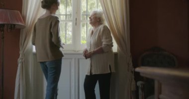 Çok nesilli kadınlar birlikte konuşuyor. Yaşlı büyükanne kadın penceresinin yakınında çay veya kahve içme ziyaret torunu ile gülümseyen. Evde beyaz saçlı yaşlı büyükanne.4k yavaş hareket