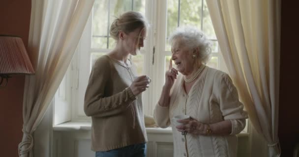 Mujeres multigeneracionales hablando juntas. Abuela anciana sonriendo con su nieta visitando cerca de la ventana bebiendo té o café.Abuela anciana de pelo blanco en casa.4k cámara lenta — Vídeos de Stock