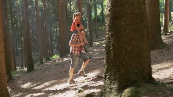Side view of father giving a piggyback ride to daughter walking in forest.Father holding daughter on shoulders hiking along forest trail path.Father and daughter hiking on forest path together — Stock Video