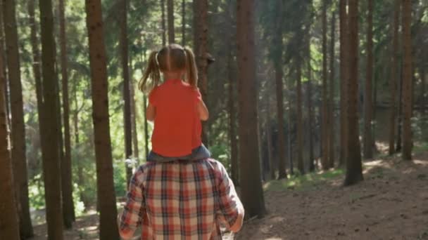 Back view of father giving a piggyback ride to daughter walking in forest.Father holding daughter on shoulders hiking along forest trail path.Father and daughter hiking on forest path together — Stock Video