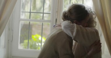 Çok nesilli kadınlar birlikte sarılmayı sever. Büyük büyükanne kadın pencereyakınında torunu veya genç arkadaşı ile mutlu. Evde beyaz saçlı yaşlı büyükanne. Sevgi,birliktelik.4k yavaş hareket