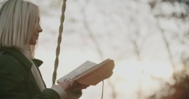 Gün batımında salıncakta kitap okuyan kadın. Gün batımında salıncakta sallanan kadın. Parkta salıncakta sallanan günlüklü bir kadın. İnsanlar günbatımında sonbahar doğasını severler. Kadın dinlenirken salıncakta sallanıyor.