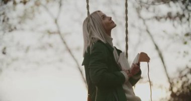 Gün batımında salıncakta kitap okuyan kadın. Gün batımında salıncakta sallanan kadın. Parkta salıncakta sallanan günlüklü bir kadın. İnsanlar günbatımında sonbahar doğasını severler. Kadın dinlenirken salıncakta sallanıyor.