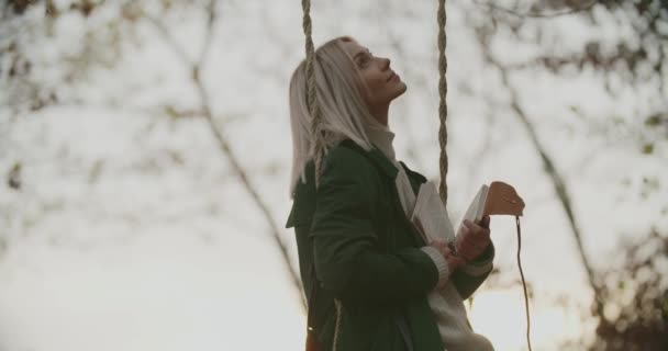 Woman reading a book on a swing at sunset.woman swinging on a swing at sunset. Woman looking diary riding a swing at park. People enjoy autumn nature at sunset.woman rides on a swing while relaxing — Stock Video