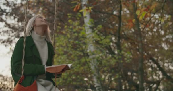 Vrouw het lezen van een boek op een schommel bij zonsondergang. vrouw swingen op een schommel bij zonsondergang. Vrouw op zoek dagboek rijden een schommel in het park. Mensen genieten van de herfst natuur bij zonsondergang.vrouw rijdt op een schommel terwijl ontspannen — Stockvideo