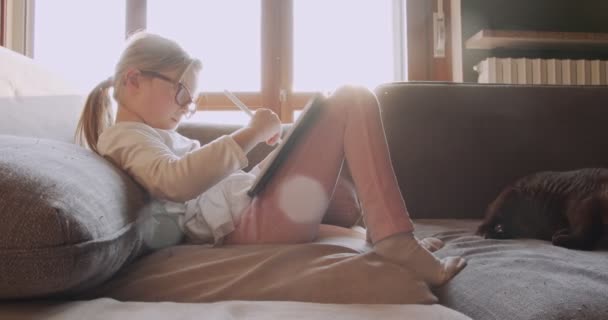 Παιδί που χρησιμοποιεί τάμπλετ στο σπίτι κάθεται στον καναπέ. Κοριτσάκι που κάθεται στον καναπέ και κάνει τα μαθήματά του με το ταμπλέτ.Θέα στο πλάι. Παιδί παίζει με δισκίο στο σαλόνι — Αρχείο Βίντεο