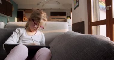 Evde tablet kullanan bir çocuk kanepede oturuyor. Küçük kız kanepede oturmuş tabletle ödev yapıyor. Çocuk oturma odasında tabletle oynuyor