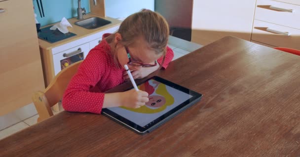 Маленька дівчинка малює на планшеті вдома. Дитячий малюнок на планшетному пристрої на кухні на дистанційній освіті. Дівчина робить домашнє завдання на планшеті. Розумна освіта. Школа вдома. Дистанційне навчання — стокове відео