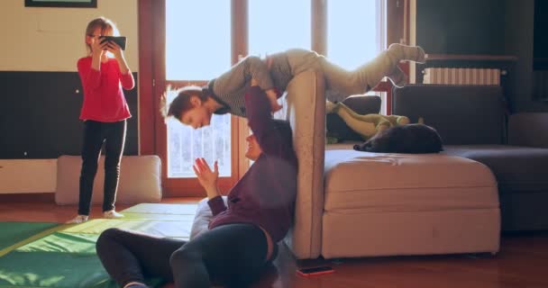 Mamá jugando con los niños juntos en casa.Mamá ayudando a su hijo a hacer un rollo, mientras que la hija de filmación en el teléfono inteligente en interiores.Vista lateral de tiro medio. Niños jugando con mamá en la sala de estar juntos.cámara lenta — Vídeo de stock