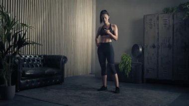 Kadın evde özel antrenör uygulaması olan akıllı telefon kullanıyor. Kadın spor yapmak için fitness uygulamasını kullanıyor. Tam yörünge çekimi. Kadın spor yaparken cep telefonu kullanıyor. 