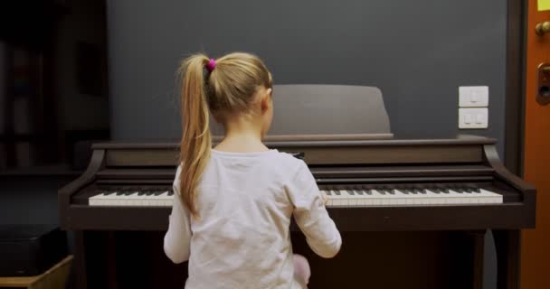 집에서 피아노치는 어린 소녀. 아이는 거실에서 피아노를 친다. 아이들은 집에서 피아노를 배우죠. 음악 수업, 중간 샷 후면 뷰. — 비디오