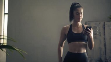 Kadın evde özel antrenör uygulaması olan akıllı telefon kullanıyor. Kadın spor yapmak için fitness uygulamasını kullanıyor. Yörüngeye yakın çekim. Kadın spor yaparken cep telefonu kullanıyor. 