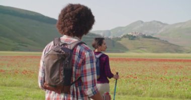 Erkek ve kadın yürüyüş direkleri kullanıyor.Castelluccio di Norcio 'daki çiçek tarlalarının yakınındaki patikada yürüyen İskandinav yürüyüşü. Güneşli bir günde Umbria' da arkadaşlarla yapılan İtalya gezisi. 60 fps el bilgisayarı.