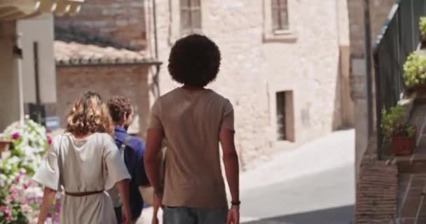 Quatro turistas felizes andando pela pequena rua visitando a cidade rural. Voltar handheld indo seguir.Grupo de pessoas multi étnicas andando na cidade velha. Férias em Italy.slow motion.Sunny tempo — Vídeo de Stock