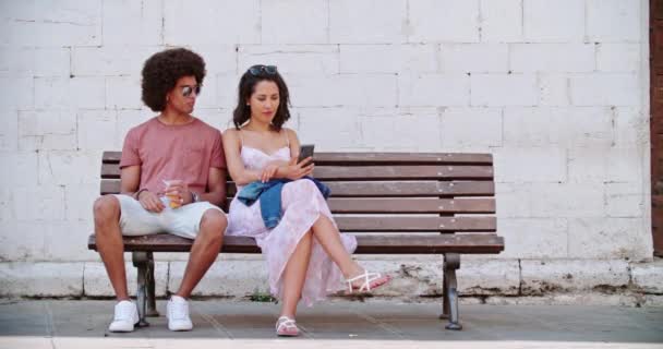 Due persone sedute su panchina che riposano utilizzando lo smartphone. Turisti che si riposano sul bench.Uomo e donna che usano il cellulare seduti sul bench.slow motion di città rurale.Viaggio tra amici in Italia. — Video Stock