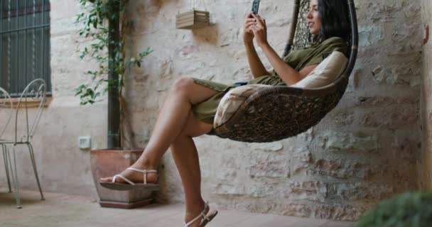 Γυναίκα χαλαρώνοντας σε καρέκλα κρέμονται χρησιμοποιώντας smartphone.Rural σπίτι. Αργή κίνηση χειροκίνητη πυροβοληθείσα πλευρική όψη.Γυναίκα κάνει μια selfie σε κρεμαστή καρέκλα. — Αρχείο Βίντεο