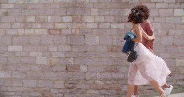 Güneşli bir günde taş duvarın önünde birlikte yürüyen iki kişi. Erkek ve kadın yürüyor ve gülümsüyor. Arka planda taş duvarla yürüyen romantik bir çift. El bilgisayarı ağır çekim kenar görünümü