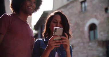 Turistler kırsal kesimdeki konforları ya da simgeleri aramak için akıllı telefon kullanıyor. Yavaş çekim. Kırsal kesimde akıllı telefon kullanırken gülümseyen iki kişi. Orta boy portre fotoğrafı. İtalya 'da arkadaş gezisi.