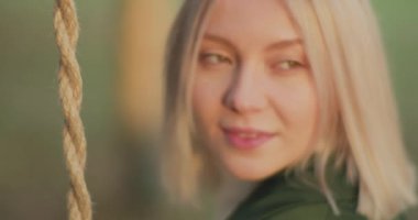 Salıncakta sallanan ve gülümseyen güzel sarışın bir kadının portresi. Sonbaharda genç bir kadının yüzünde gün batımı ışığı. Ağır çekim.