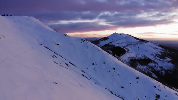 Flygfoto över snötäckta bergssluttning på vintern under soluppgång eller solnedgång. Panorama av hög snö bergskedja, i bakgrunden rosa färgade moln. slow motion etablering sköt antenn bergssidan — Stockvideo