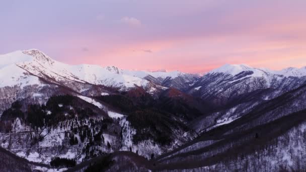 Flygfoto över snötäckta bergskedjor under rosa och gul solnedgång eller soluppgång. Mörk skog i dalen. slow motion upprättande skott — Stockvideo