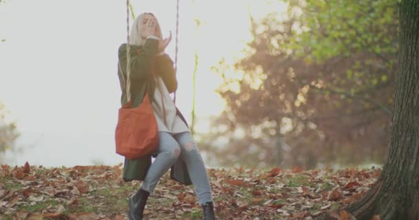 Mladá žena pije horký čaj, dívá se na západ slunce na zádech na houpačce pod velkým stromem v chladném podzimním cestě.zpomalení — Stock video