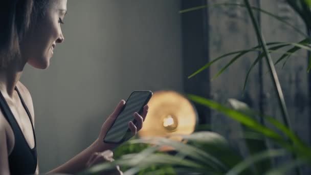 Boční pohled na mladé fitness a brunetky žena se usmívá při pohledu na mobil během cvičení odpočinku v obývacím pokoji doma — Stock video