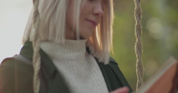 Mulher lendo um livro sobre um balanço ao ar livre.Memórias positivas ressurgir para uma mulher jovem e bonita, enquanto balançando e segurando um livro, no outono. Movimento lento. — Vídeo de Stock