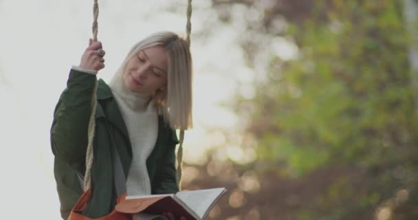 Young woman reads a book. Beautiful woman swinging and smiles looking in the distance. slow motion. sun backlight at sunset — Stock Video