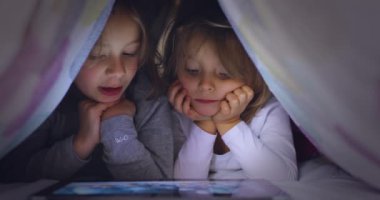 Arkadaşı ya da kız kardeşi olan çocuk sarışın beyaz kız geceleri yatak örtüsünün altında tablet uygulamasında film ya da animasyon izliyor. Modern teknoloji çocukluk çocuğu evde kullanıyor. 4k video
