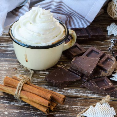 Krem şantili Enamel kupa sıcak çikolata, bir dilim acı çikolata ve ahşap arka planda tarçın çubukları. Kış zamanları içki konsepti. Lezzetli soğuk hava içeceği.