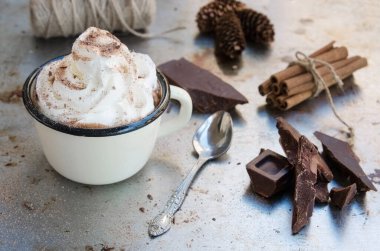 Krem şantili Enamel kupa sıcak çikolata, bir dilim acı çikolata ve metal vintage arka planda tarçın çubukları. Kış zamanları içki konsepti. Lezzetli soğuk hava içeceği.